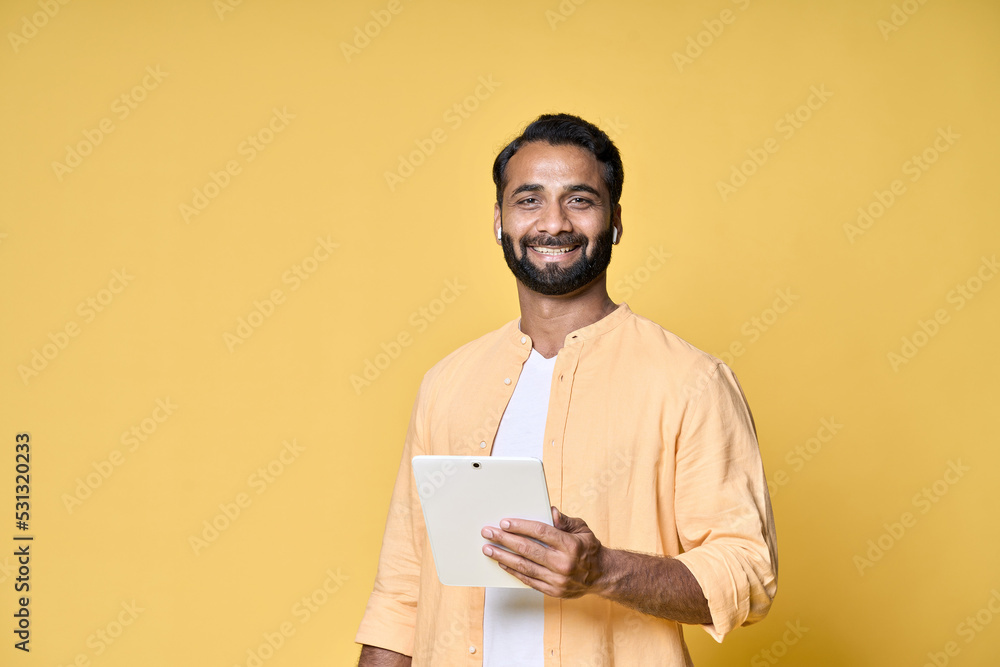 Happy indian man using digital tablet standing isolated on yellow background. Smiling ethnic guy holding pad wearing earbud advertising virtual class webinars or audio books. Happy indian man using digital tablet standing isolated on yellow background. Smiling ethnic guy holding pad wearing earbud advertising virtual class webinars or audio books.