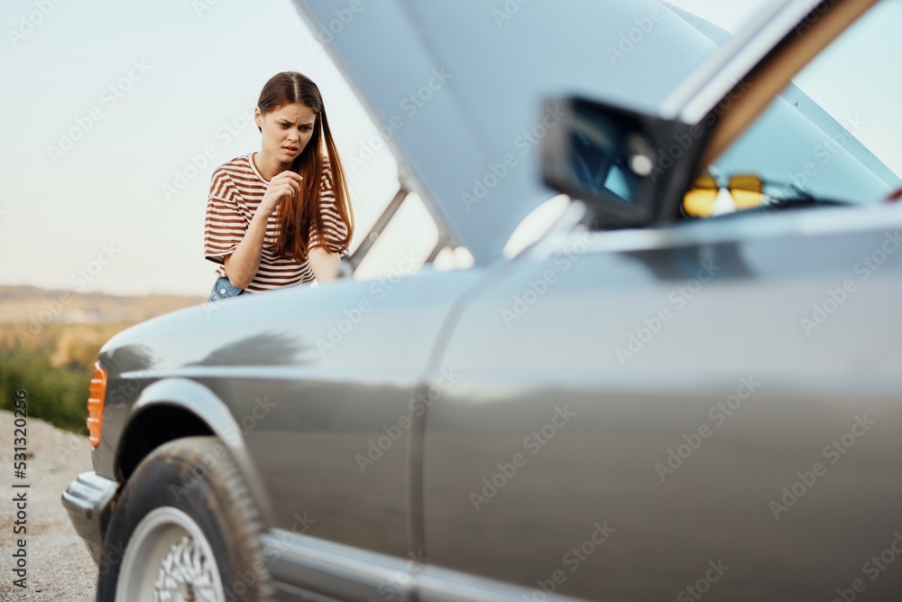 A woman traveler stands by her old car with the hood open, looking for the cause of the car breakdown alone without men on the road in the countryside