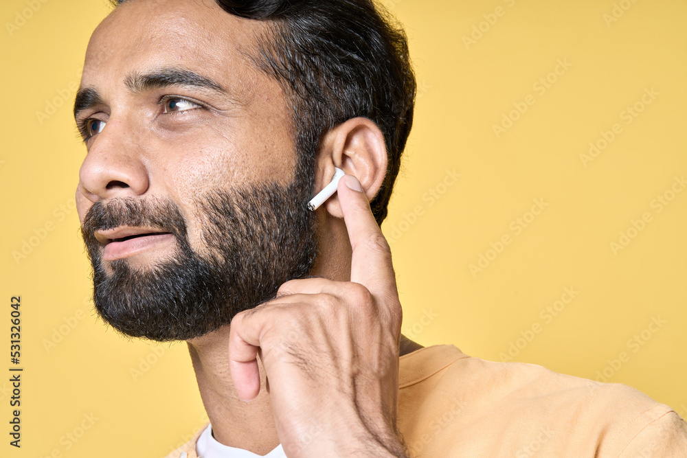 Fototapeta premium Thoughtful smiling bearded indian man relaxing wearing earbud thinking while listening to music, business podcast or audio book in mobile application isolated on yellow background. Close up portrait