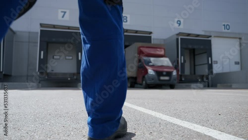 Engineer walks towards truck waiting for unloading at large storage terminal. Large industrial building with open gates backside view slow motion