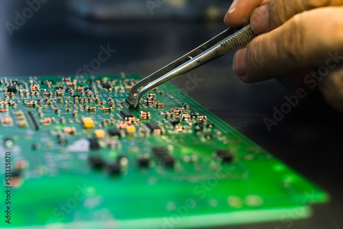 Close-up shot of PCB printed circuit board with mounted electrical components and hand with tweezers replacing parts. Repairing electronics. Precision and focus. Horizontal shot. High quality photo