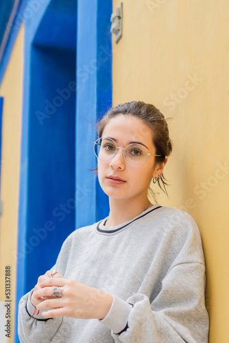 Woman wearing glasses and looking at camera leaning on a yellow wall.