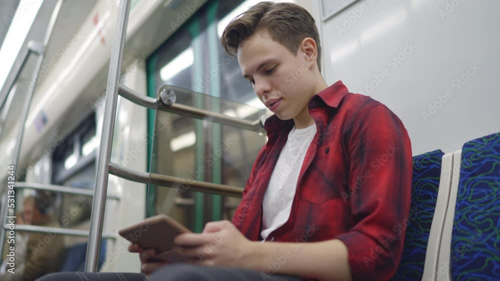 A young man in a checkered shirt sits on a subway train and looks at something on a pocket computer. Subway, computer tablet, model, student. Slow motion 120fps footage A young man in a checkered shirt sits on a subway train and looks at something on a pocket computer. Subway, computer tablet, model, student. Slow motion 120fps footage