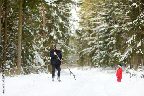 Cute toddler boy and his granddad having fun on a walk in snow covered pine forest on chilly winter day.