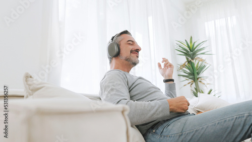 Attractive Bearded mature adult man sitting on the couch playing an air guitar and listening to music with headphones, Lifestyle concept, Relax in living room at home.