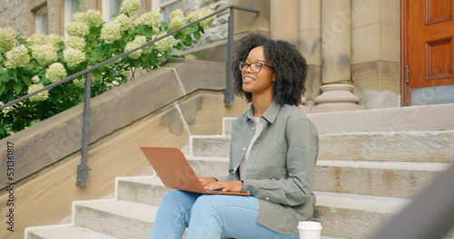 African American young female, seating outdoors on the stairs, she works with her lap top computer, and takes a moment to look a side for inspiring thought