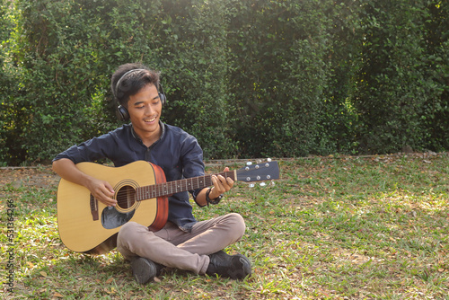 Portrait of attractive Asian man in casual shirt playing guitar sitting on garden grass