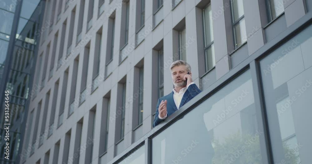 Bottom View of Caucasian Businessman Standing at the Street with Office Center at the Background. Senior Professional Talking on Phone. Outside Bottom View of Caucasian Businessman Standing at the Street with Office Center at the Background. Senior Professional Talking on Phone. Outside
