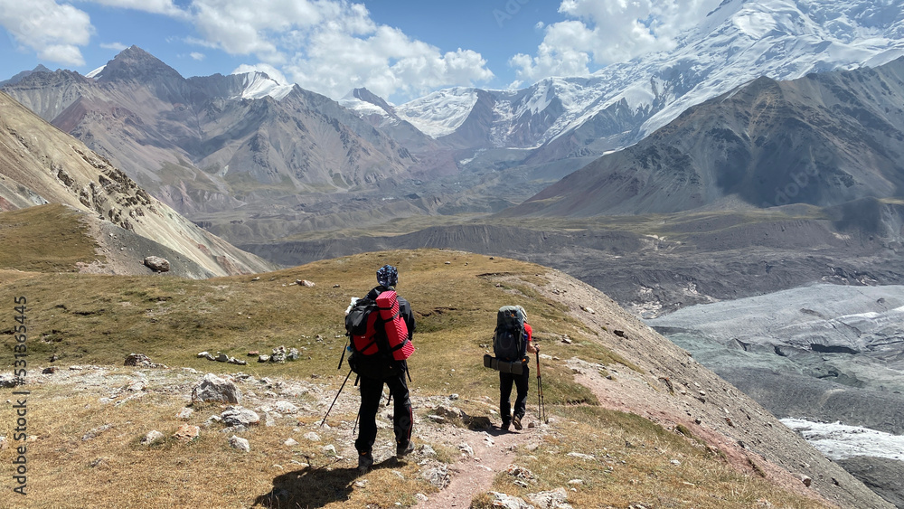 Two tourists with big backpacks are walking in the mountains. View of the rocky mountains, snow-capped peaks and hills of Kyrgyzstan.