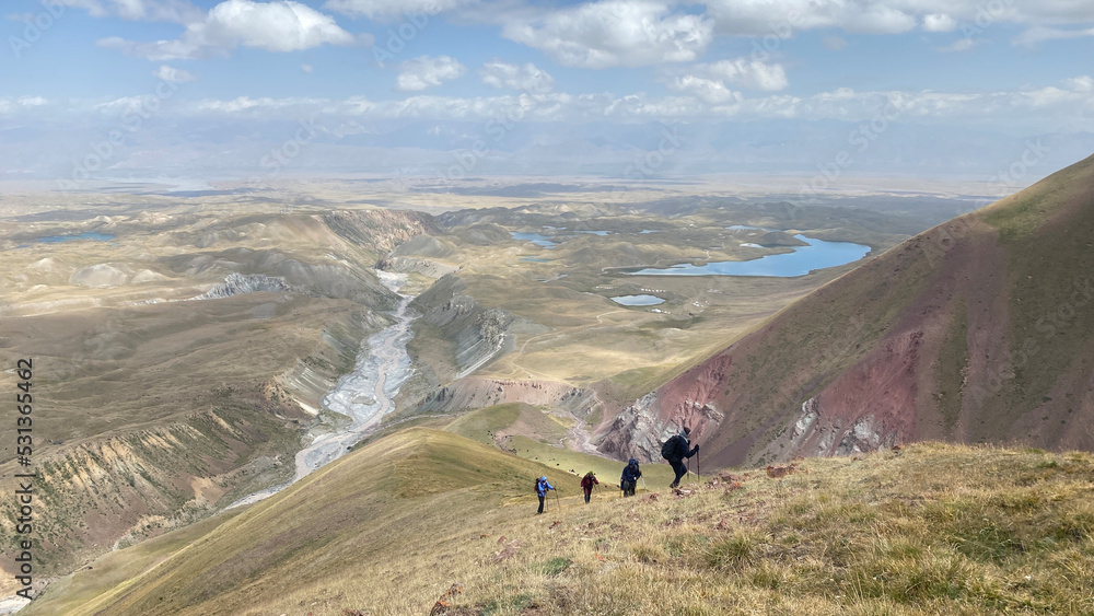 A group of tourists goes up the mountain. View from a height of the Alai Valley. Colored rocky mountains, lakes, rivers, green slopes and hills of Kyrgyzstan.