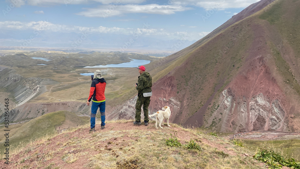 Two tourists with a dog stand on a high hill, look into the distance and take a photo on the phone. View from a height of colorful rocky mountains, lakes, green slopes and hills of Kyrgyzstan. 