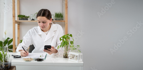 Tableau sur toile A young attractive woman scientist with protective gloves using a smartphone in the scientific bio laboratory, writing results on a clipboard