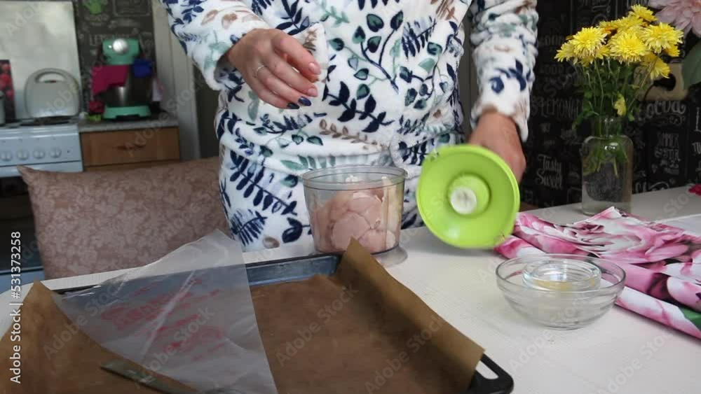 A woman pours spices into a blender bowl with chicken fillet. Starts to
