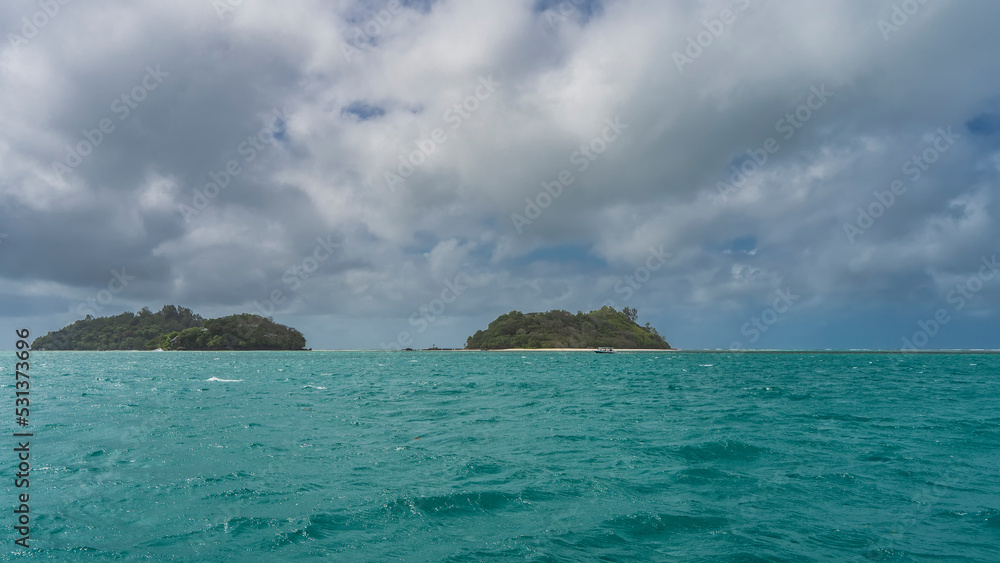 Two tropical islands are completely overgrown with green vegetation. A sandy beach is visible. The boat sails on the turquoise ocean. Blue sky with picturesque clouds. Seychelles