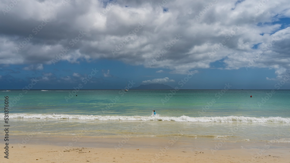 Beautiful tropical beach. The waves of the turquoise ocean roll ashore. A man is swimming in the foam of the waves. The ship in the distance. The silhouette of the island against the blue sky, clouds.