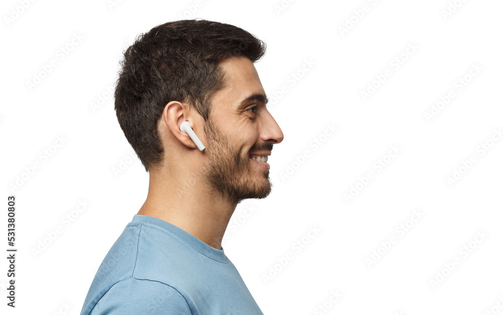 Fototapeta premium Sideways portrait of smiling young man listening to music or radio, uses modern wireless earphones, wearing blue t-shirt