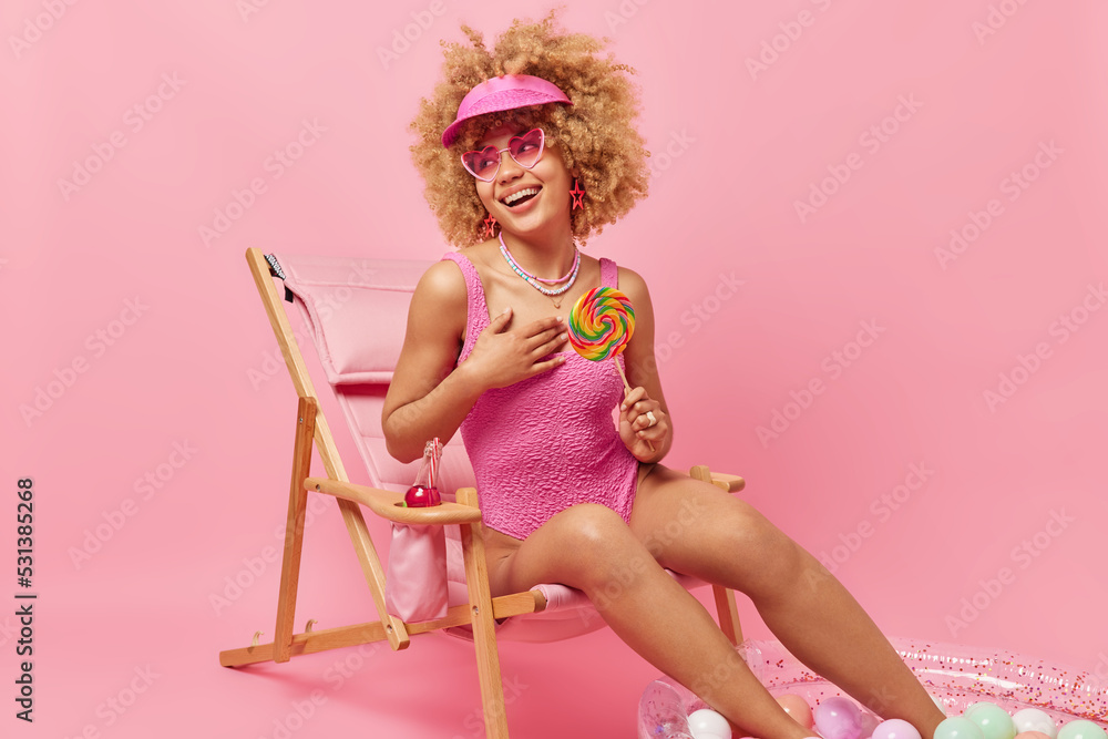Happy curly haired woman wears panama sunglasses and swimsuit holds big multicolored lollipop looks happily away enjoys summer vacation and sunbathing poses on lounge chair against pink wall Happy curly haired woman wears panama sunglasses and swimsuit holds big multicolored lollipop looks happily away enjoys summer vacation and sunbathing poses on lounge chair against pink wall