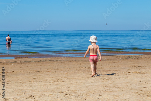 A two-year-old Toddler boy walks towards the sea alone in swimming trunks and panama