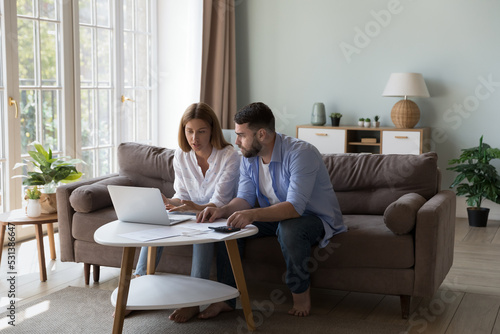 Serious millennial husband and wife discussing family expenses, using online payment laptop, talking, doing accounting job, paperwork, calculating budget, mortgage, rent fees