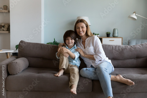 Happy loving mom hugging cute preschool son, looking at camera, smiling, enjoying motherhood. Cheerful kid and mother resting on couch. Family leisure concept. Home portrait