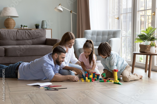 Family couple of happy parents and two little son and daughter kids playing learning games on warm floor together, building towers from colorful toy construction blocks, enjoying leisure, playtime