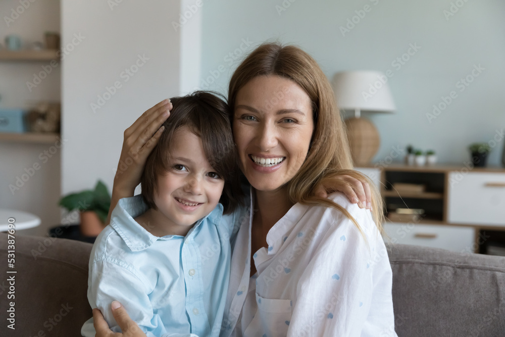 Cheerful loving mother and little son kid hugging on couch, looking at camera, smiling. Mother embracing cute preschool child with care, affection. Motherhood concept. Family head shot portrait Cheerful loving mother and little son kid hugging on couch, looking at camera, smiling. Mother embracing cute preschool child with care, affection. Motherhood concept. Family head shot portrait