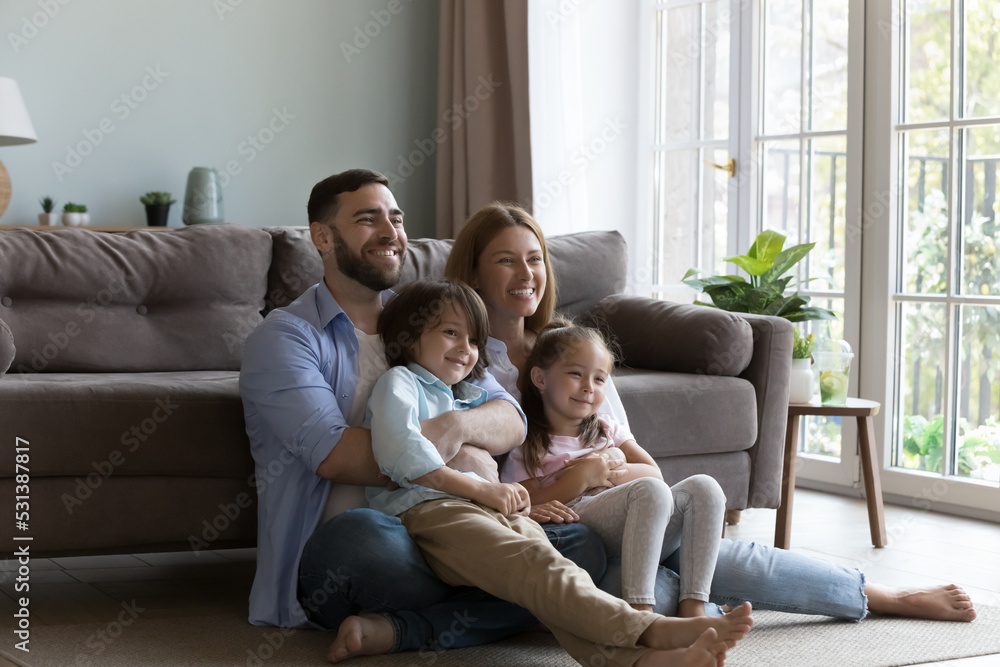 Cheerful Caucasian parents and cute sibling boy and girl resting on heating floor together, laughing, looking away, watching TV, posing for shooting, enjoying leisure. Family home portrait