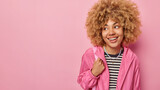 Studio shot of good looking teenage girl with curly hair look away gladfully smiles happily dressed in striped jumper and windbreaker isolated over pink background empty space for your text.
