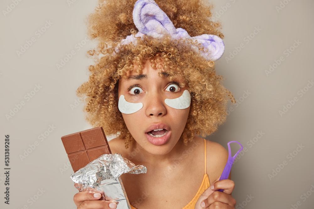 Headshot of amazed scared woman with bushy blonde hair holds tongue scraper and bar of chocolate stares with omg expression applies beauty patches to reduce wrinkles poses against grey background - obrazy, fototapety, plakaty 