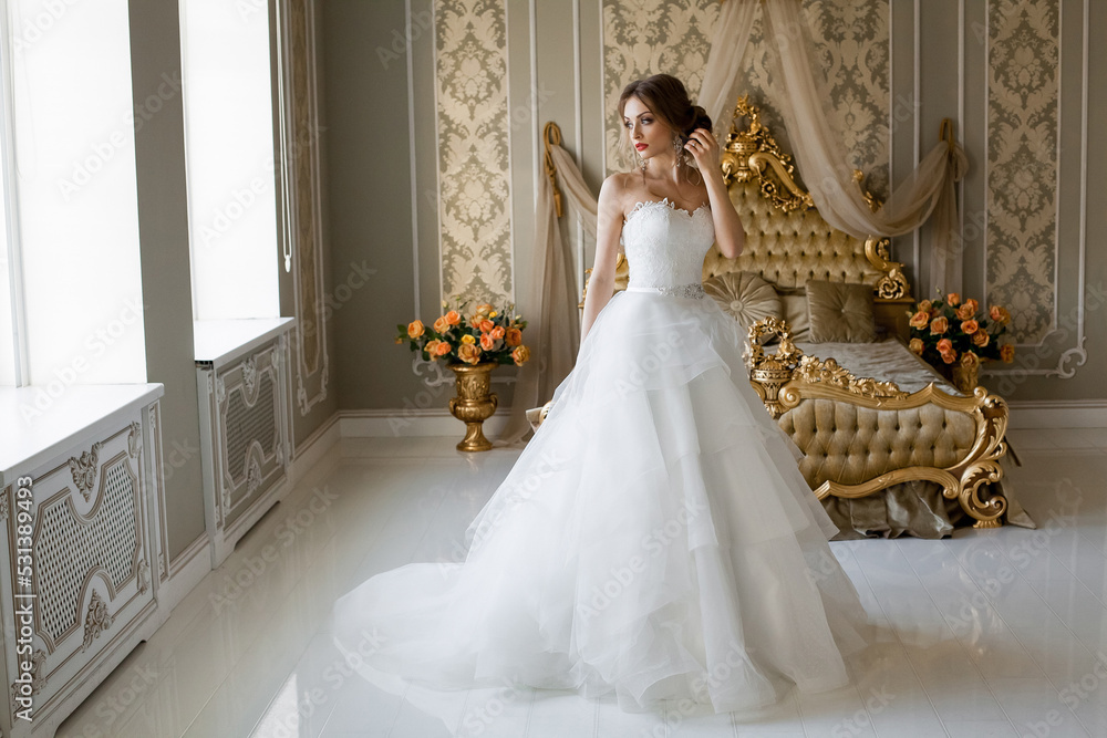 A beautiful smiling bride with chic makeup and hairstyle in a white wedding dress is standing in a bedroom with a stylish light vintage interior, in the morning before the wedding. A beautiful smiling bride with chic makeup and hairstyle in a white wedding dress is standing in a bedroom with a stylish light vintage interior, in the morning before the wedding.