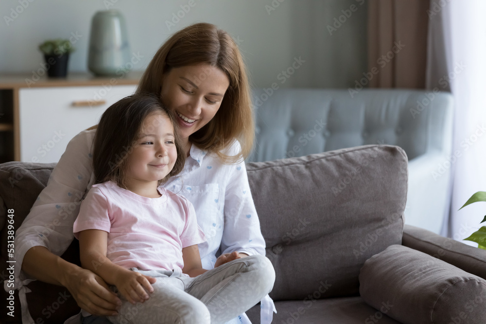 Cheerful mom holding happy cute little daughter in arms on sofa, cuddling kid with love, care, talking to thoughtful dreamy girl looking away. Motherhood, family leisure concept Cheerful mom holding happy cute little daughter in arms on sofa, cuddling kid with love, care, talking to thoughtful dreamy girl looking away. Motherhood, family leisure concept