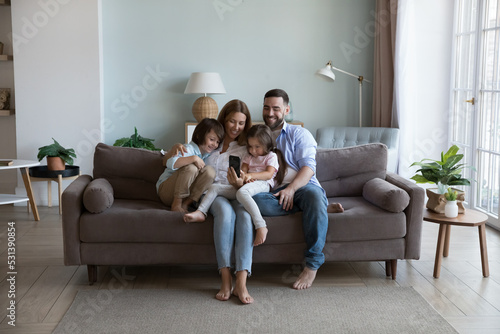 Quadro su tela Happy millennial parents and two kids using mobile phone, resting on home couch together, holding smartphone, taking selfie, relaxing in living room interior