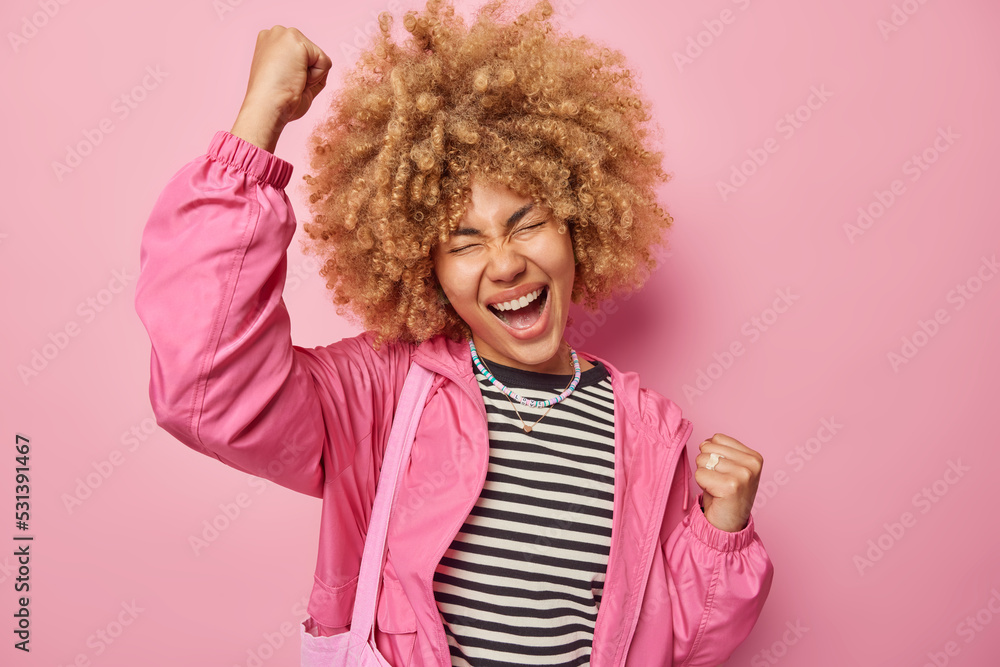 Overjoyed curly haired female model does winner gesture exclaims loudly celebrates success dressed in casual clothes feels ambitious and encouraged makes fist bump isolated over pink background Overjoyed curly haired female model does winner gesture exclaims loudly celebrates success dressed in casual clothes feels ambitious and encouraged makes fist bump isolated over pink background