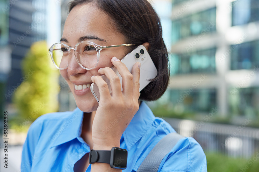 Outdoor shot of beautiful young Asian woman with dark hair has telephone call talks with friend enjoys positive conversation phones via app wears transparent eyeglasses blue shirt and smartwach Outdoor shot of beautiful young Asian woman with dark hair has telephone call talks with friend enjoys positive conversation phones via app wears transparent eyeglasses blue shirt and smartwach
