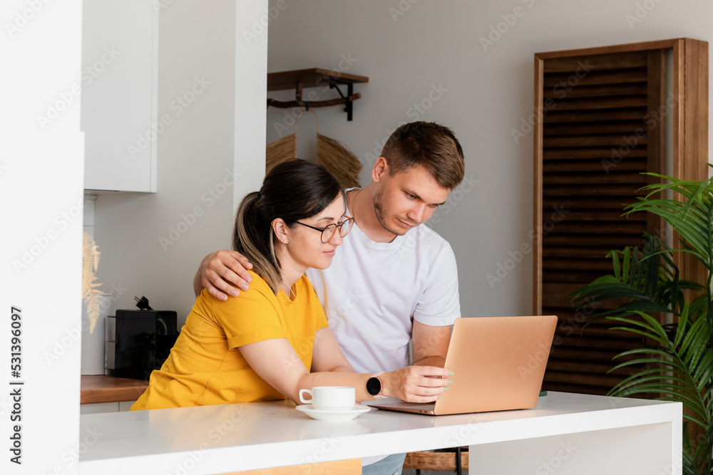 Pleasant married couple is sitting in a modern kitchen looking at a laptop screen. A young family is surfing the web, shopping online or booking flights. Pleasant married couple is sitting in a modern kitchen looking at a laptop screen. A young family is surfing the web, shopping online or booking flights.