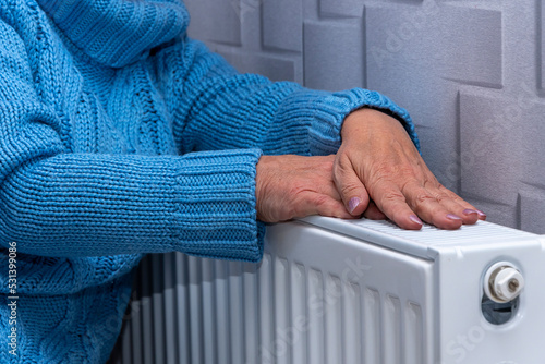 An elderly woman in a blue sweater warming her hands on a heating radiator. Concept: low room temperature, rising gas and heating prices, getting colder in the apartment. photo