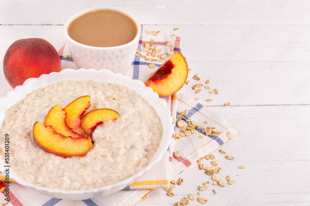 Oatmeal porridge with peach in a bowl (plate) and coffee in a cup on a white wooden background. Place for text, recipe. Healthy breakfast.