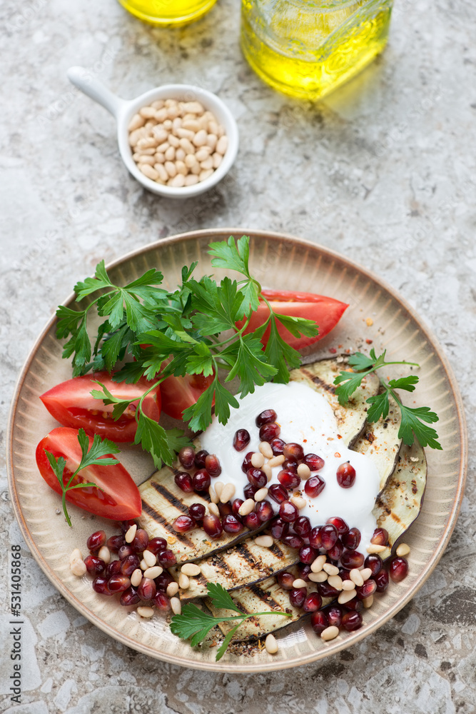 Grilled slices of aubergine with pomegranate, pine nuts, yogurt, tomatoes and parsley, elevated view on a light-brown granite background, vertical shot Grilled slices of aubergine with pomegranate, pine nuts, yogurt, tomatoes and parsley, elevated view on a light-brown granite background, vertical shot