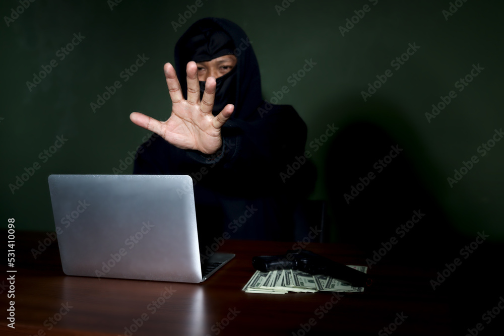 Criminal man in black hood with laptop computer, a gun and money at the at desk reaching out his hand to camera , mask thief robber committing committed cyber crimes online. Criminal man in black hood with laptop computer, a gun and money at the at desk reaching out his hand to camera , mask thief robber committing committed cyber crimes online.