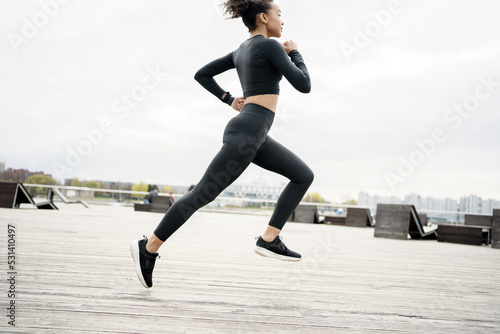 A woman in a tight tracksuit and sneakers trains outside. A fitness watch and a running app are on hand.
