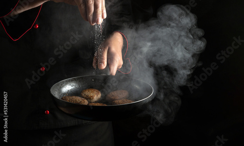 Fototapeta Naklejka Na Ścianę i Meble -  Professional chef adds salt to a hot pan with meat burger cutlets. Free space for hotel menu or recipe on black background