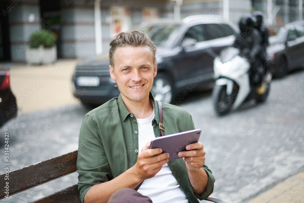 Portrait of young freelancer man work using digital tablet sits on the bench outdoors in front office building wearing casual green shirt and shoulder bag. Young handsome man working on the go Portrait of young freelancer man work using digital tablet sits on the bench outdoors in front office building wearing casual green shirt and shoulder bag. Young handsome man working on the go