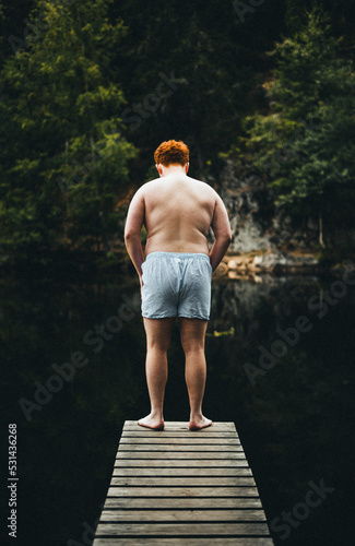 Rothaariger übergewichtiger Junge steht auf einem Steg im Wald und überlegt in das Wasser im See zu springen