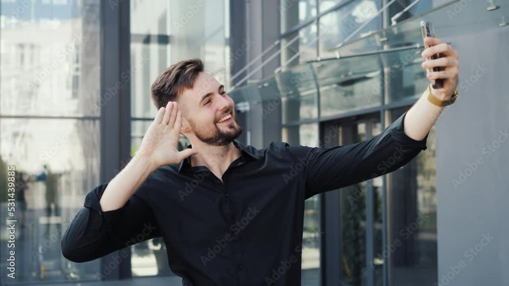 Smiling young business man making selfie picture by phone video call standing outdoor at business district. Portrait of a businessman blogger shooting video blog outdoors near modern office building. Smiling young business man making selfie picture by phone video call standing outdoor at business district. Portrait of a businessman blogger shooting video blog outdoors near modern office building.