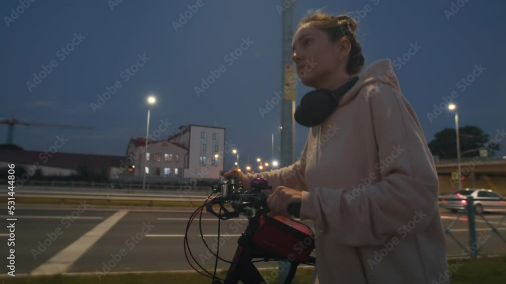 Young female in casual sport clothes walks along the street by night and riding her bike. Woman come back home after her daily routine evening stroll, city nightlife with illuminated street lights Young female in casual sport clothes walks along the street by night and riding her bike. Woman come back home after her daily routine evening stroll, city nightlife with illuminated street lights