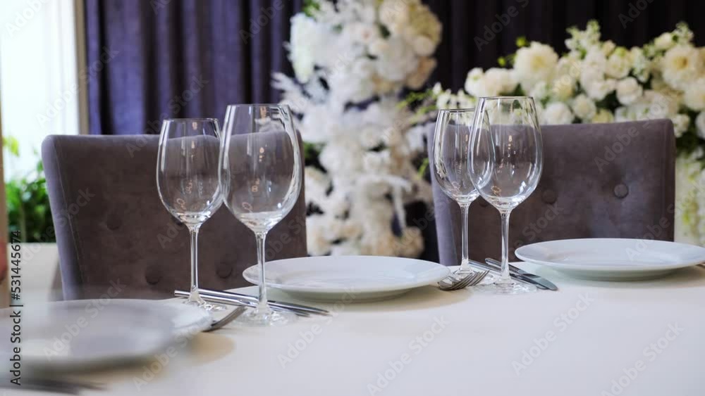 Clean and empty sets of two glasses stand near plates on banquet restaurant table. Stylish light serving background with beautiful flowers. Tablecloth catering, celebration, festive, light, party. Clean and empty sets of two glasses stand near plates on banquet restaurant table. Stylish light serving background with beautiful flowers. Tablecloth catering, celebration, festive, light, party.
