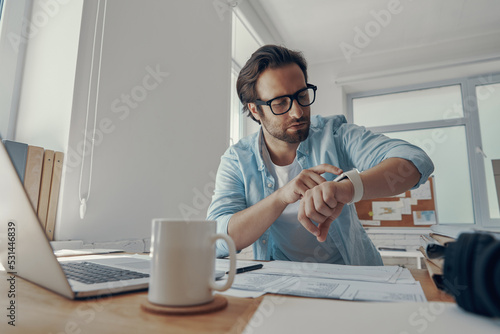 Confident young man looking at his smart watch while sitting at his working place in office