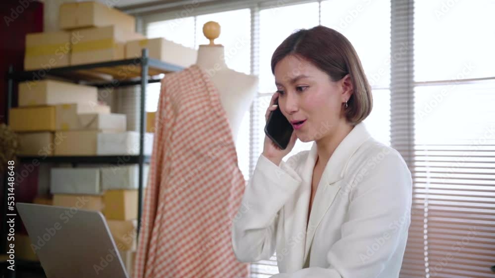 Beautiful asian entrepreneur woman call phone with consulting client distantly in tailoring atelier, discuss order. Successful fashion designer have online smartphone with consumer at work shop.
