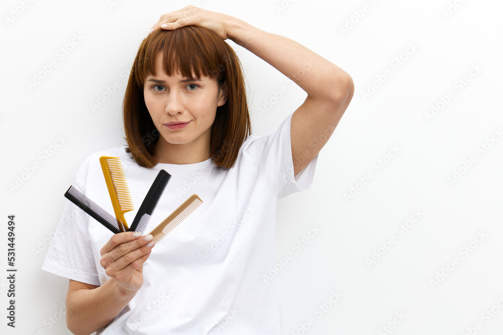 a sweet, pleasant woman with a beautiful hairstyle holds a lot of different combs in one hand, and touches her head with the other, standing in a white T-shirt on a white background a sweet, pleasant woman with a beautiful hairstyle holds a lot of different combs in one hand, and touches her head with the other, standing in a white T-shirt on a white background