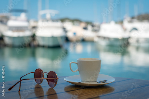 White cup with saucer, sunglasses on a table on the background of blurred boat harbor. Summer vacation concept photo